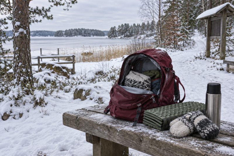 Vinröd dagsryggsäck öppen på en torr träbänk vid en snötäckt stigstart i låglandet, med sittunderlag i skum, tumvantar och en stängd termos bredvid